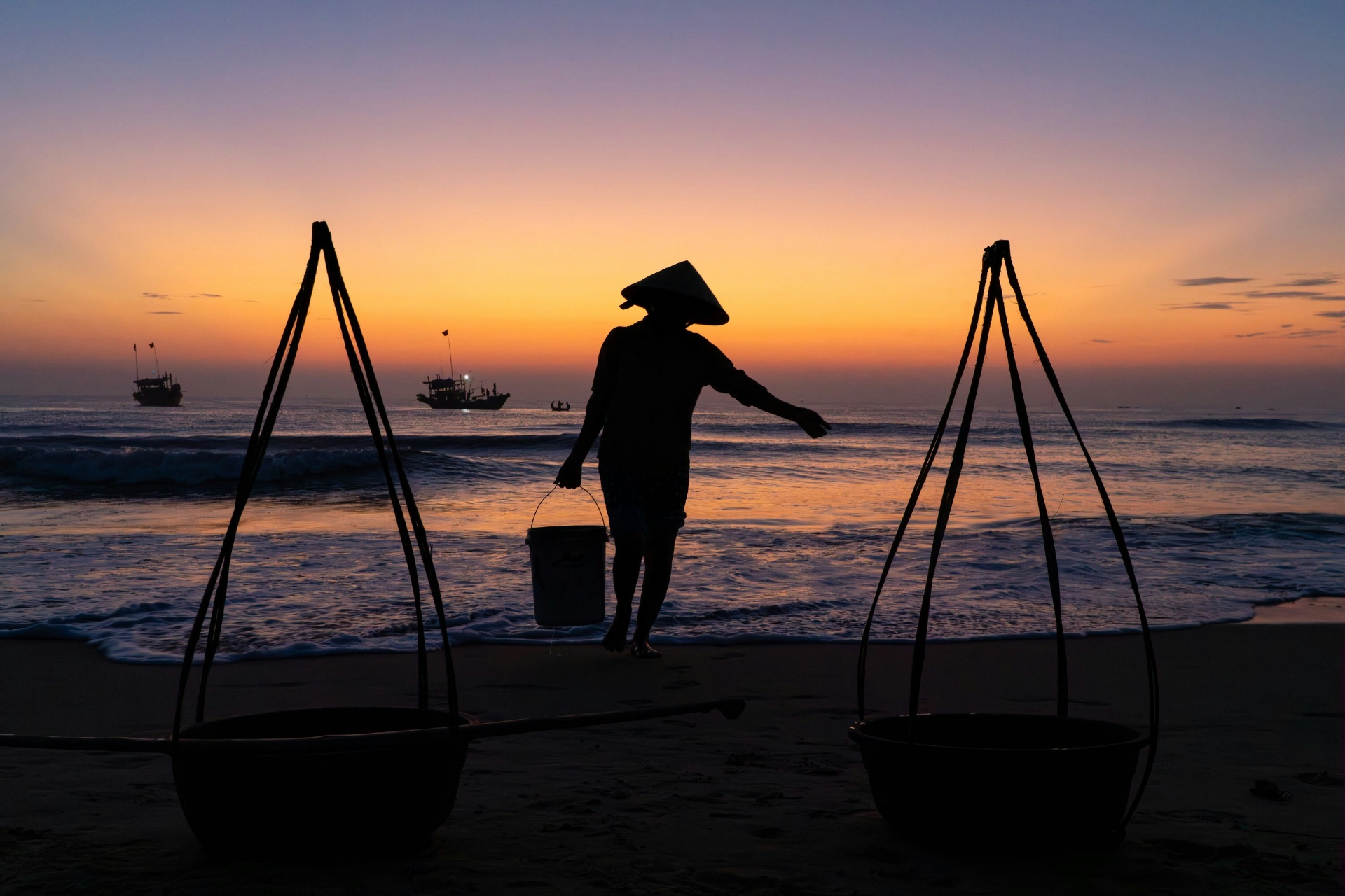 A local fisherwoman in a conical hat carrying a bucket along the shoreline at sunset, with fishing boats on the sea and woven baskets on the sand